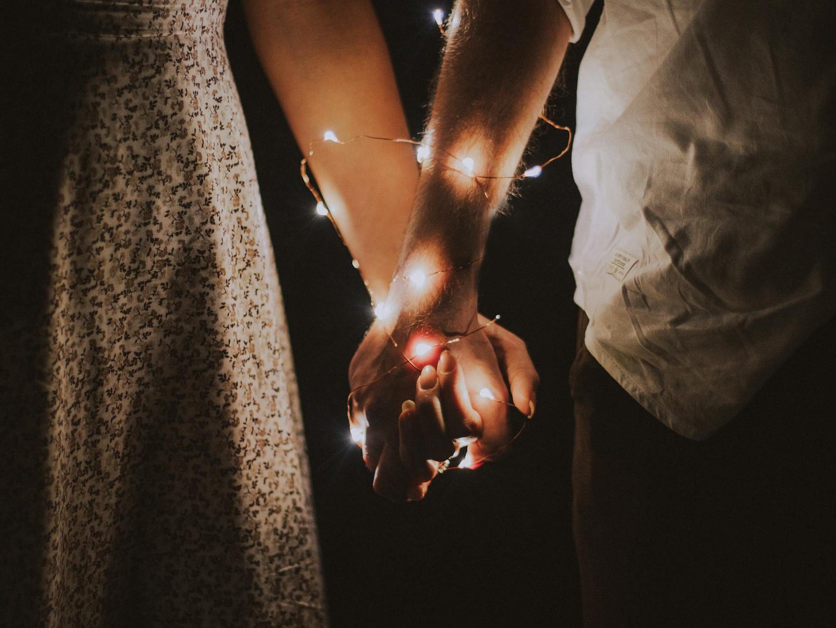 A warm and intimate close-up of a couple holding hands adorned with glowing string lights.